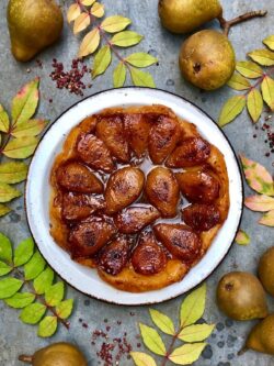 A white dish with a tarte tatin made of large brown juicy pear halves in a brown sauce on a puffy pastry.
