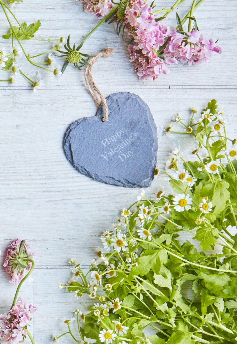 A personalised hanging heart slate on a wooden ground and a bunch of fresh daisy flowers below it.