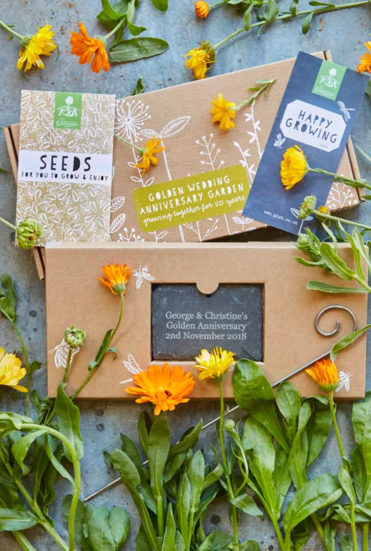 A decorated cardboard presentation box with a golden wedding anniversary garden label and a packet of seeds, a hanging slate in a cardboard box, a gift card and yellow and orange calendula flowers decoratively spread around it.