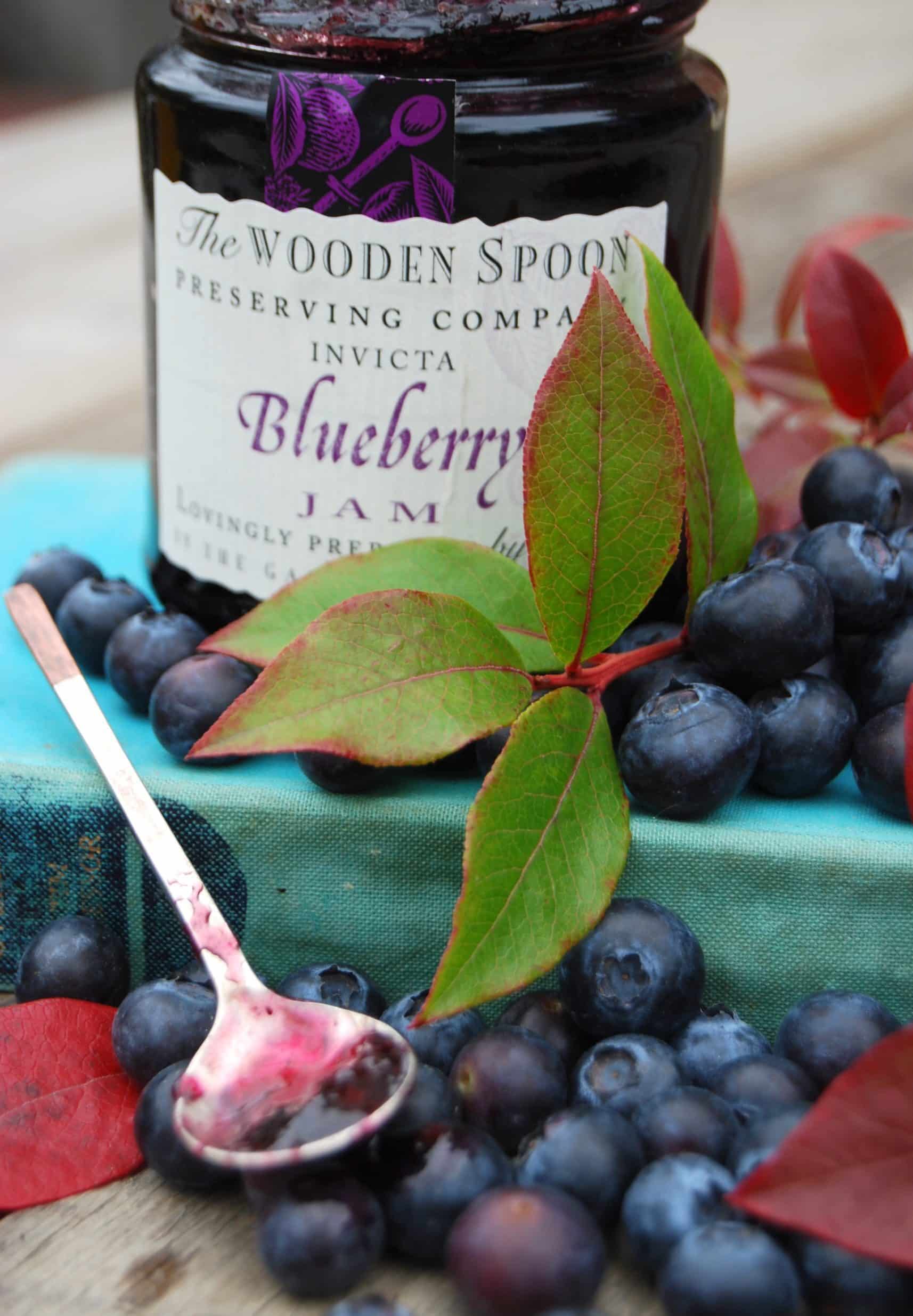 A jar of blueberry jam surrounded by fresh blueberries, a green blueberry leaf and a small spoon filled with blueberry jam.