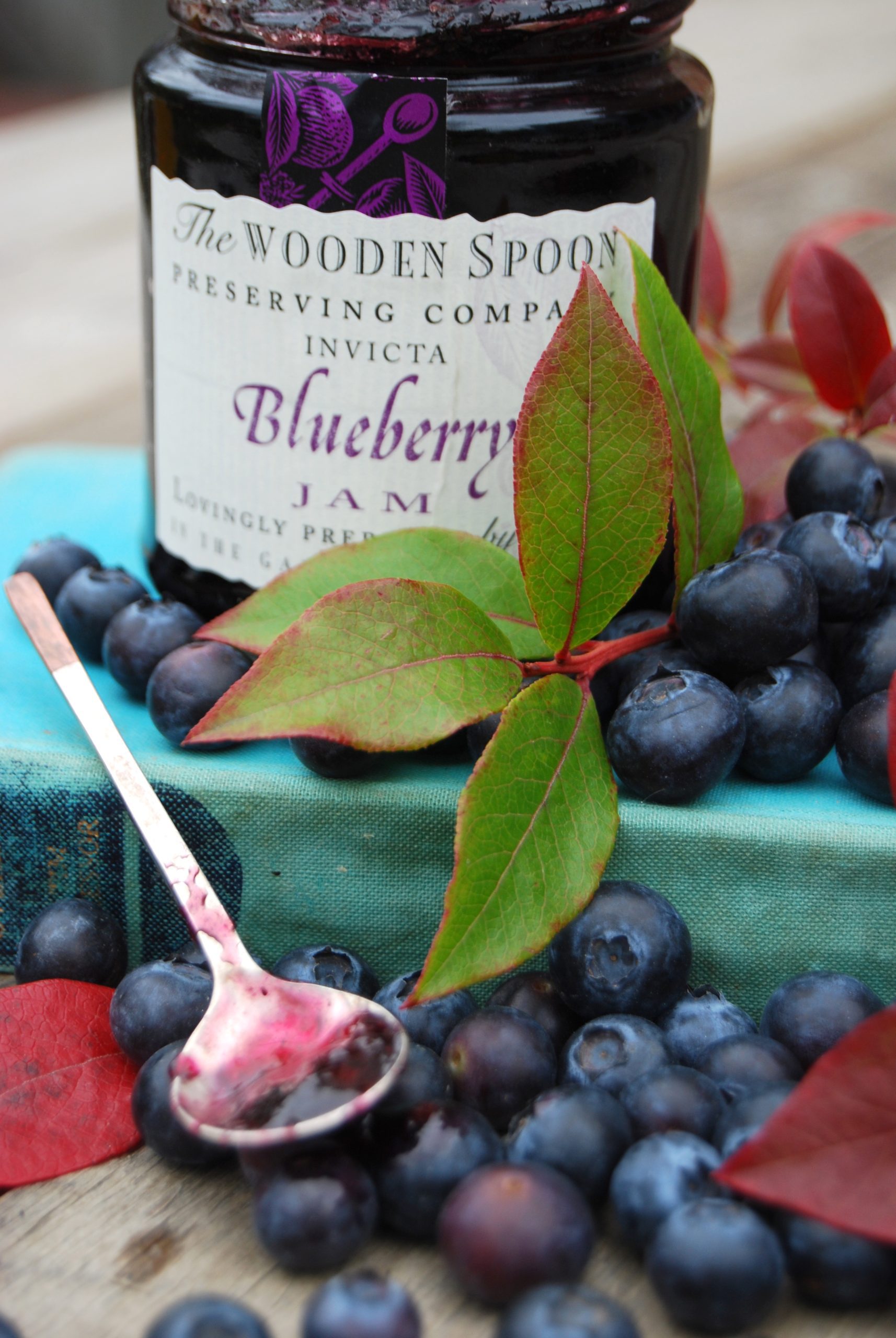 A jar of blueberry jam surrounded by fresh blueberries, a green blueberry leaf and a small spoon filled with blueberry jam.