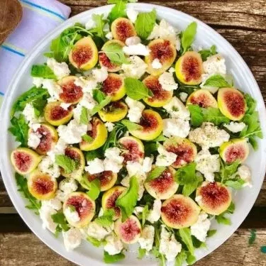 A large white plate with a Salad containing fig halves, mint leaves and soft goats cheese on a wooden table next to a napkin with two wooden salad spoons.