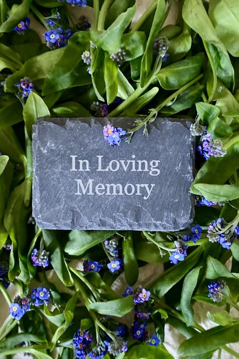 A small personalised engraved slate nestled in Forget-me-nots with bright green leaves and small blue flowers.