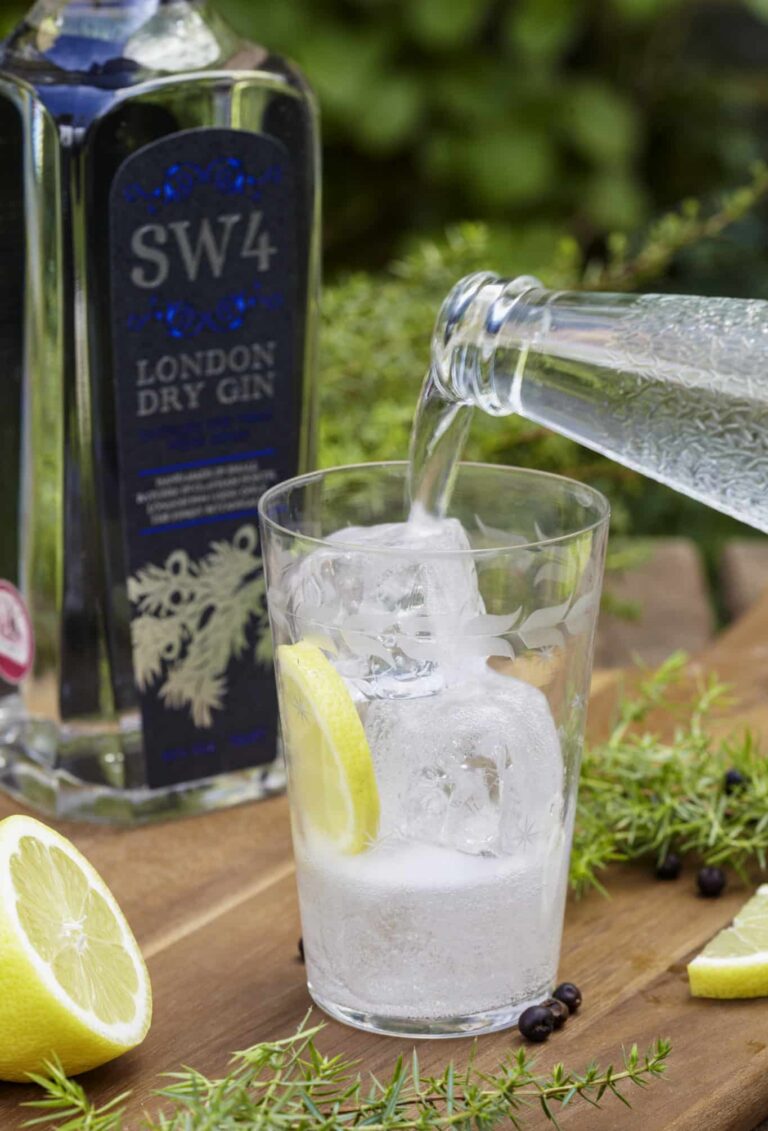 A glass filled with filled with 1/3rd gin, ice cubes and a slice of lemon with water being poured into it, a bottle of dry gin standing in the background, together with half a lemon, a few juniper twigs and juniper berries spread around the glass.