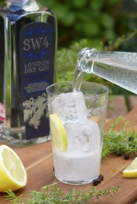 A glass filled with filled with 1/3rd gin, ice cubes and a slice of lemon with water being poured into it, a bottle of dry gin standing in the background, together with half a lemon, a few juniper twigs and juniper berries spread around the glass.