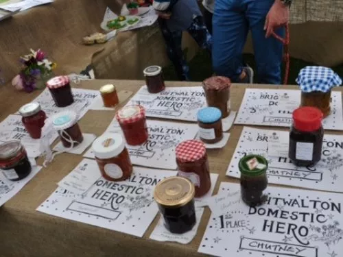 A table with a diverse selection of jars with chutney, jam and molasses placed on 'domestic heroes' flyers.