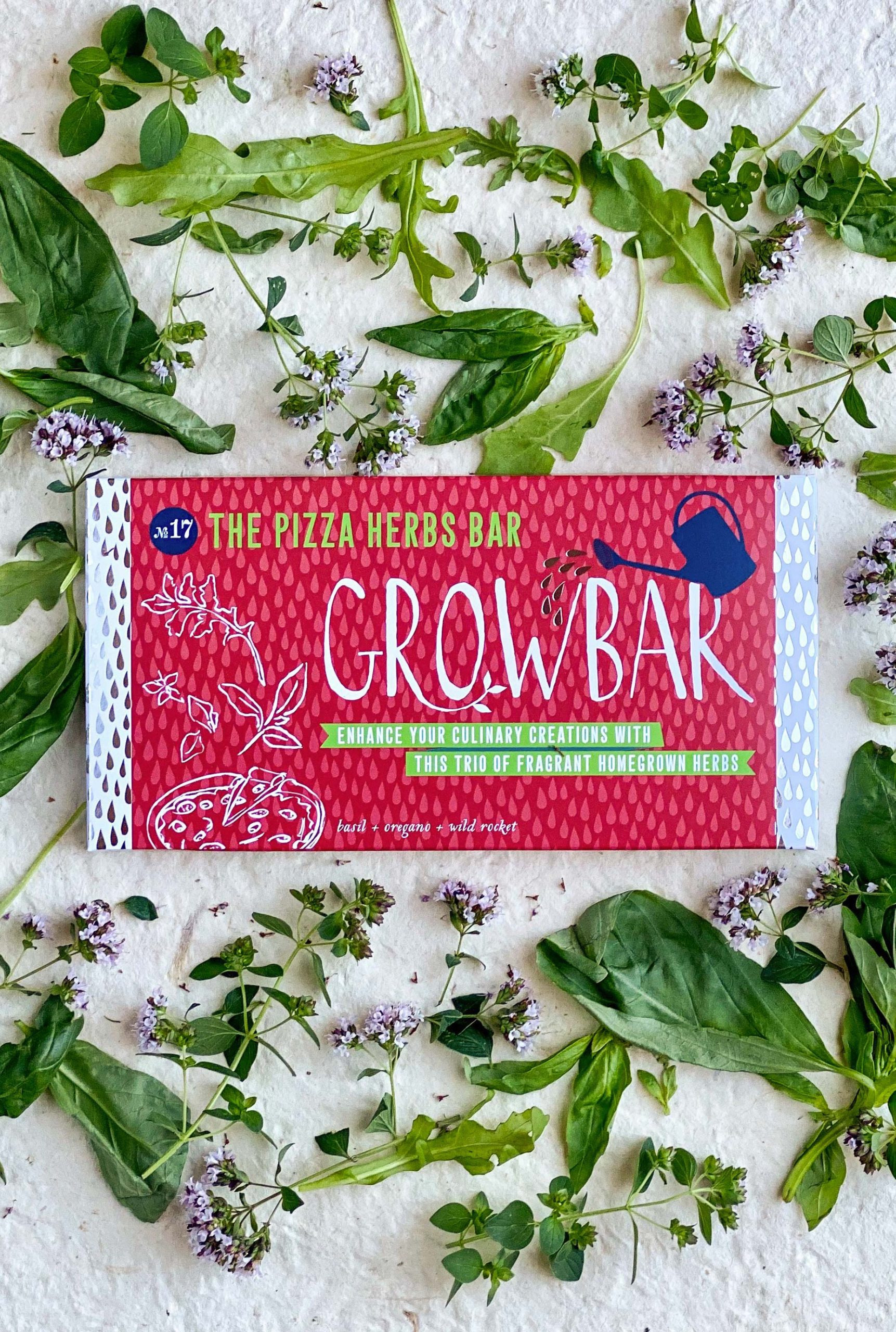 A red illustrated grow bar package surrounded by a wild arrangement of basil leaves, wild rocket and flowering oregano.