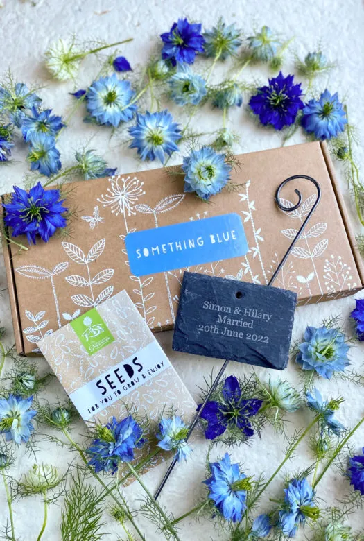 A decorated cardboard presentation box with a 'something blue' label, a personalised hanging slate, a packet of seeds and dried 'love -in-a-mist' flowers in royal blue and bright blue colour.