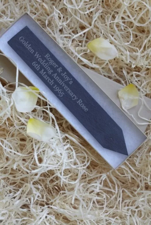 A personalised engraved slate plant stake reading 'Roger & Joy's Golden Wedding Anniversary Rose 6th March 1965' in an opened box. on a bed of wood wool surrounded by four yellow rose petals.