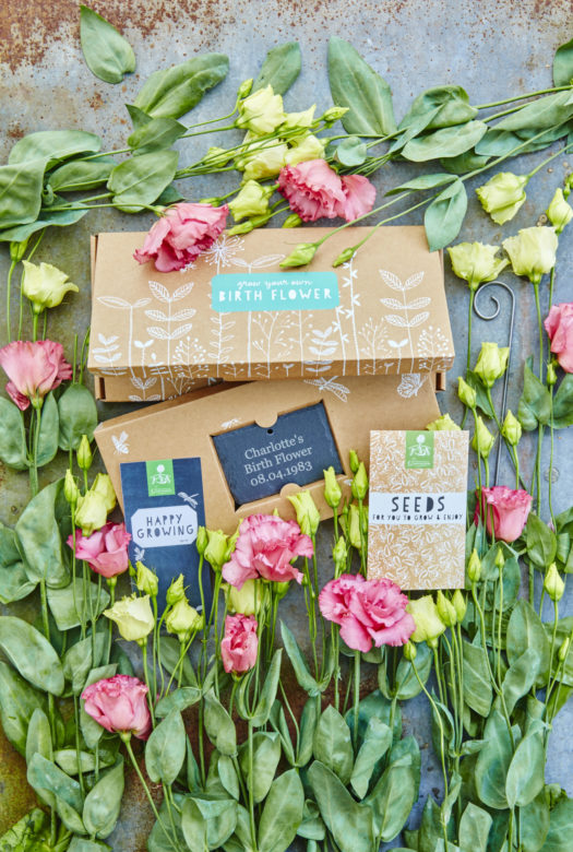 A beautifully decorated cardboard gift box for growing birth flowers with a personalised engraved garden slate. a packet of seeds and a gift card with arrangement of pink and white Lisianthus flowers decoratively framing the gift set.