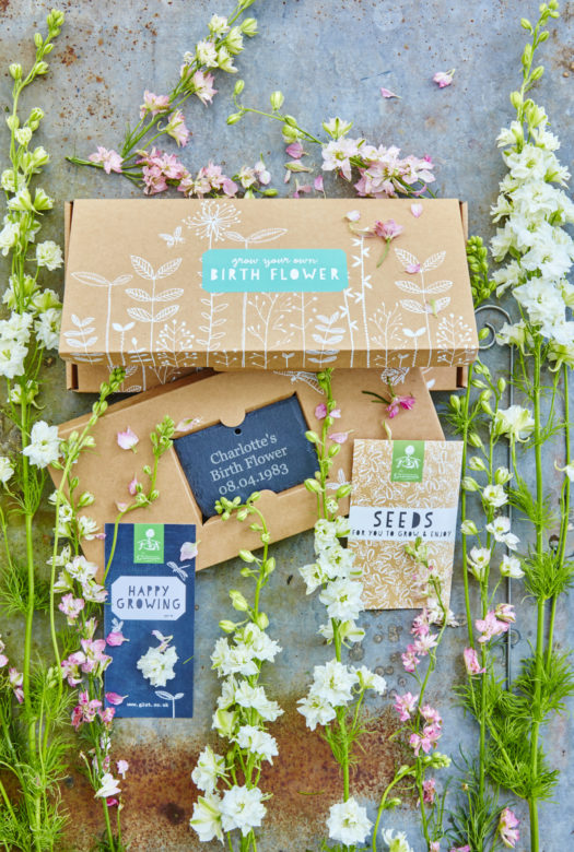 A beautifully decorated cardboard gift box for growing birth flowers with a personalised engraved garden slate. a packet of seeds and a gift card with arrangement of white and pink Larkspur flowers decoratively framing the gift set.