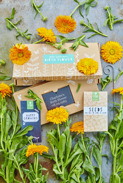 A beautifully decorated cardboard gift box for growing birth flowers with a personalised engraved garden slate. a packet of seeds and a gift card with arrangement of orange Marigold flowers decoratively framing the gift set.