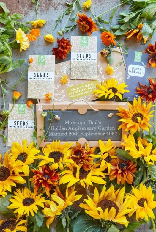 A selection of three packets of seeds on a decorated cardboard presentation box, next to a gift card and a large personalised garden slate. At the bottom a rich bouquet of bright yellow sunflowers and a few loose golden Cone Flowers and Marigold flowers spread around the box.