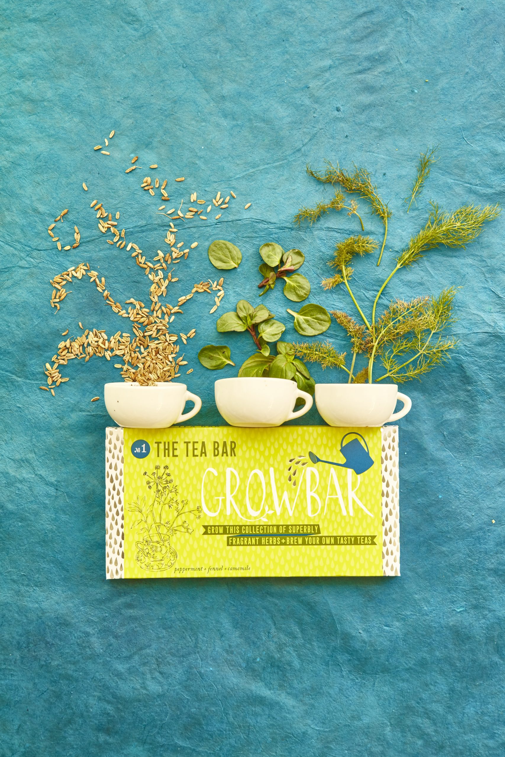 A green tea growbar with three small white teacups and chamomile seeds, peppermint leaves and fennel leaves placed above.