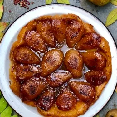 A white dish with a tarte tatin made of large brown juicy pear halves in a brown sauce on a puffy pastry.