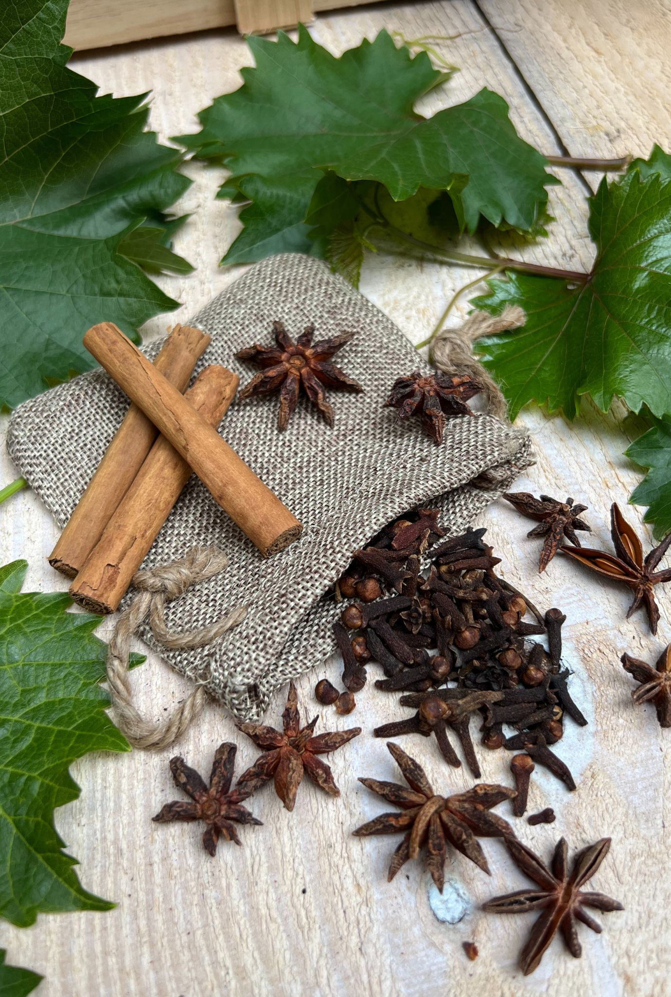 Close up of plant leaves, star anise, cinnamon sticks and cloves in hessian bag on wooden flooring.