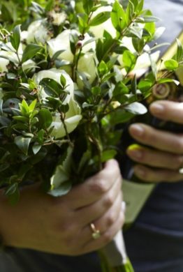 A bouquet of green leafy myrtle twigs and white roses held by a hand with a bottle of champagne held by the other hand.