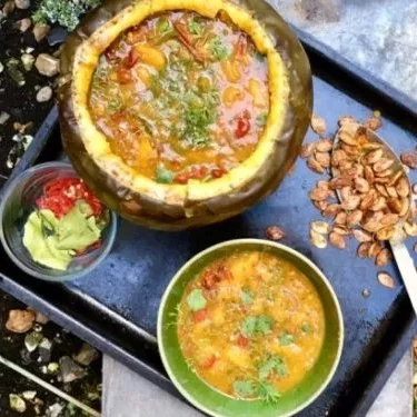 A black tray holding an ensemble of a halved pumpkin containing a spiced pumpkin laksa soup, a small bowl with pumpkin soup and coriander leaves, a large spoon filled with pumpkin seeds and a small glass dish filled containing avocado and red sliced chillies.