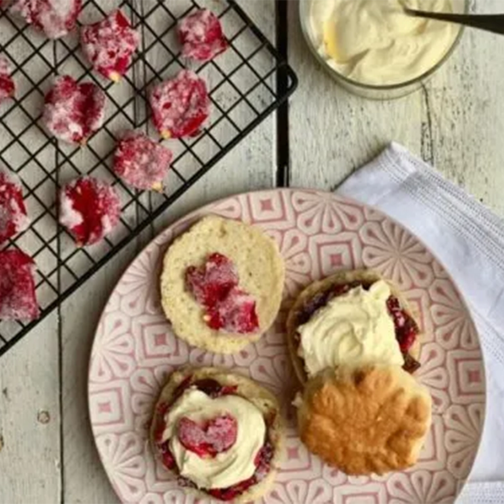 A wooden table set with a rose coloured plate with two scones, halved and spread with cream cheese, jam and candied rose petals, places alongside a white cotton napkin and a tray with sugared edible rose petals.