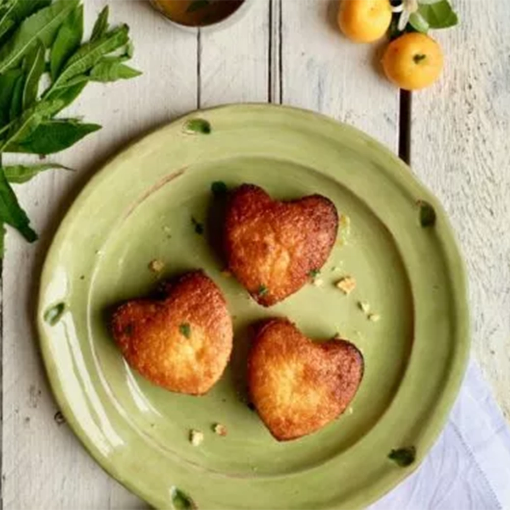 A green plate on a wooden table with three small heart shaped Calamondin Orange and Almond cakes next to a bunch of green leaves and small yellow fruits.