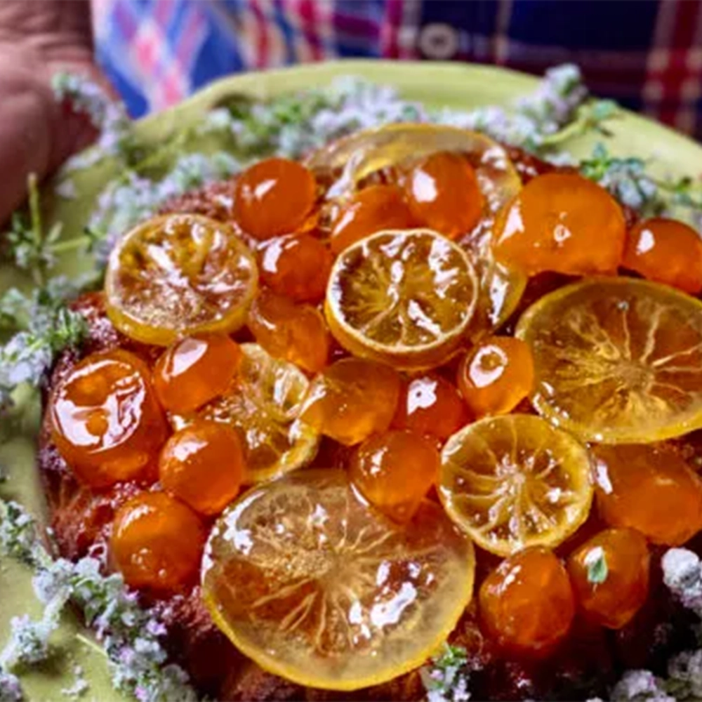 An orange fruit cake with jewel like sugared Calamondin oranges lemon slices on a green dish surrounded by sugared flowers.