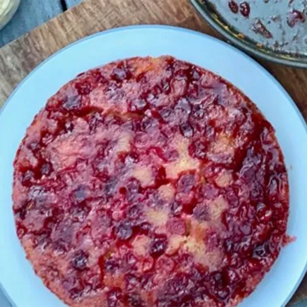 A cranberry cake with ruby red juicy cranberries on a white plate on top of a wooden board and a small knife.