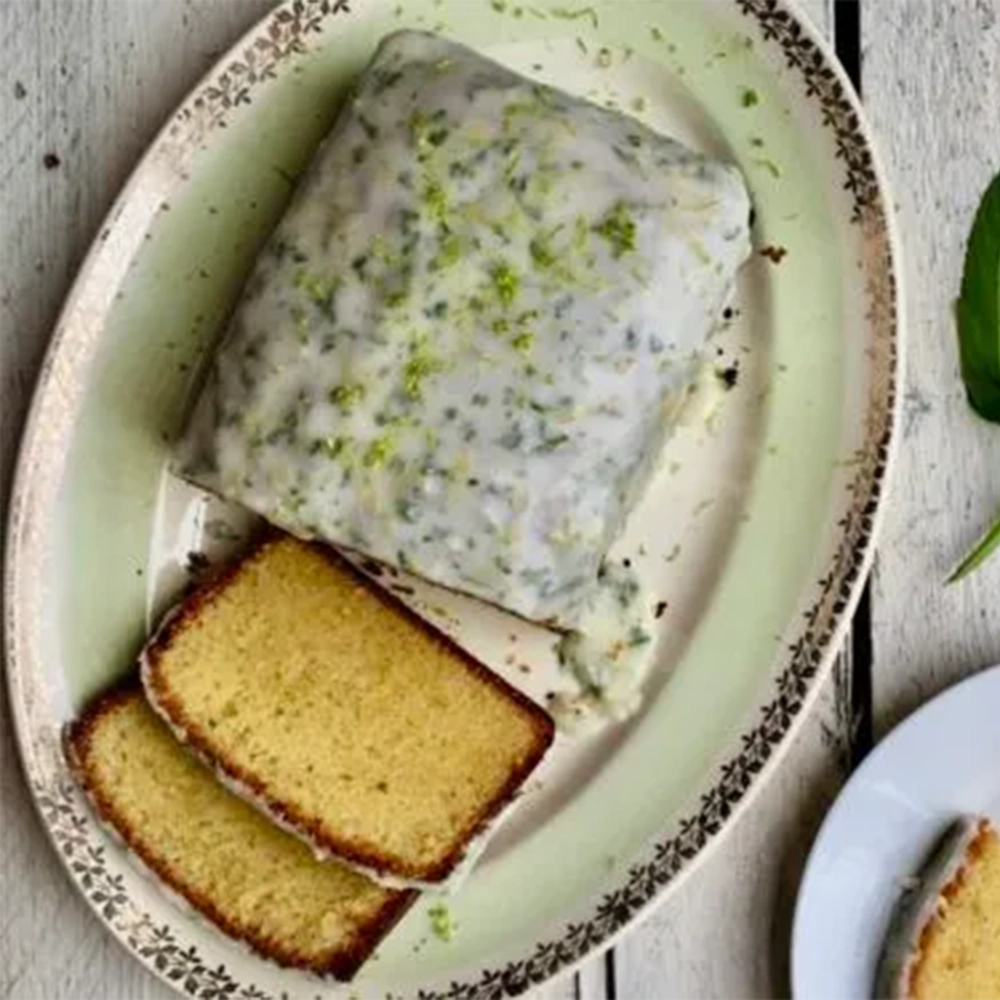 A sliced gin and lime loaf cake with a sugar frosting, lime zest and chopped peppermint leaves on a mint green decorative cake plate with a twig of peppermint leaves.