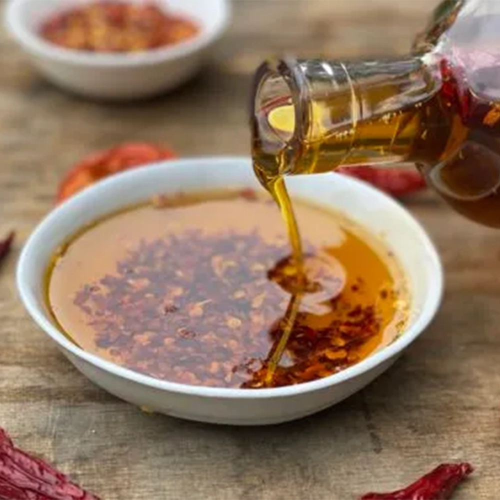 A small deep filled with a red coloured Chilli Oil and chilli flakes that is being poured from a glass bottle. Some dried chillies are placed around the dish.