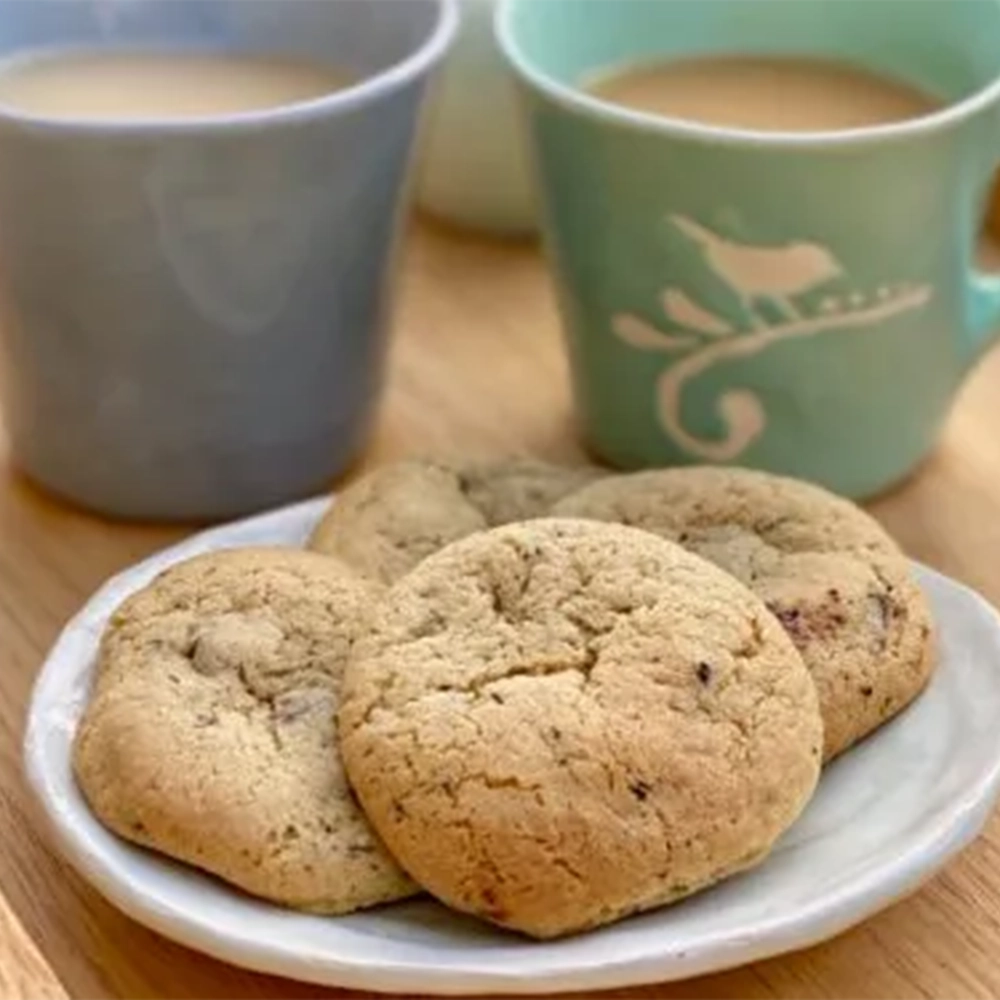 A small ceramic plate with four round chocolate chip cookies/ biscuits paired with two cups of black tea with milk on a wooden tray.