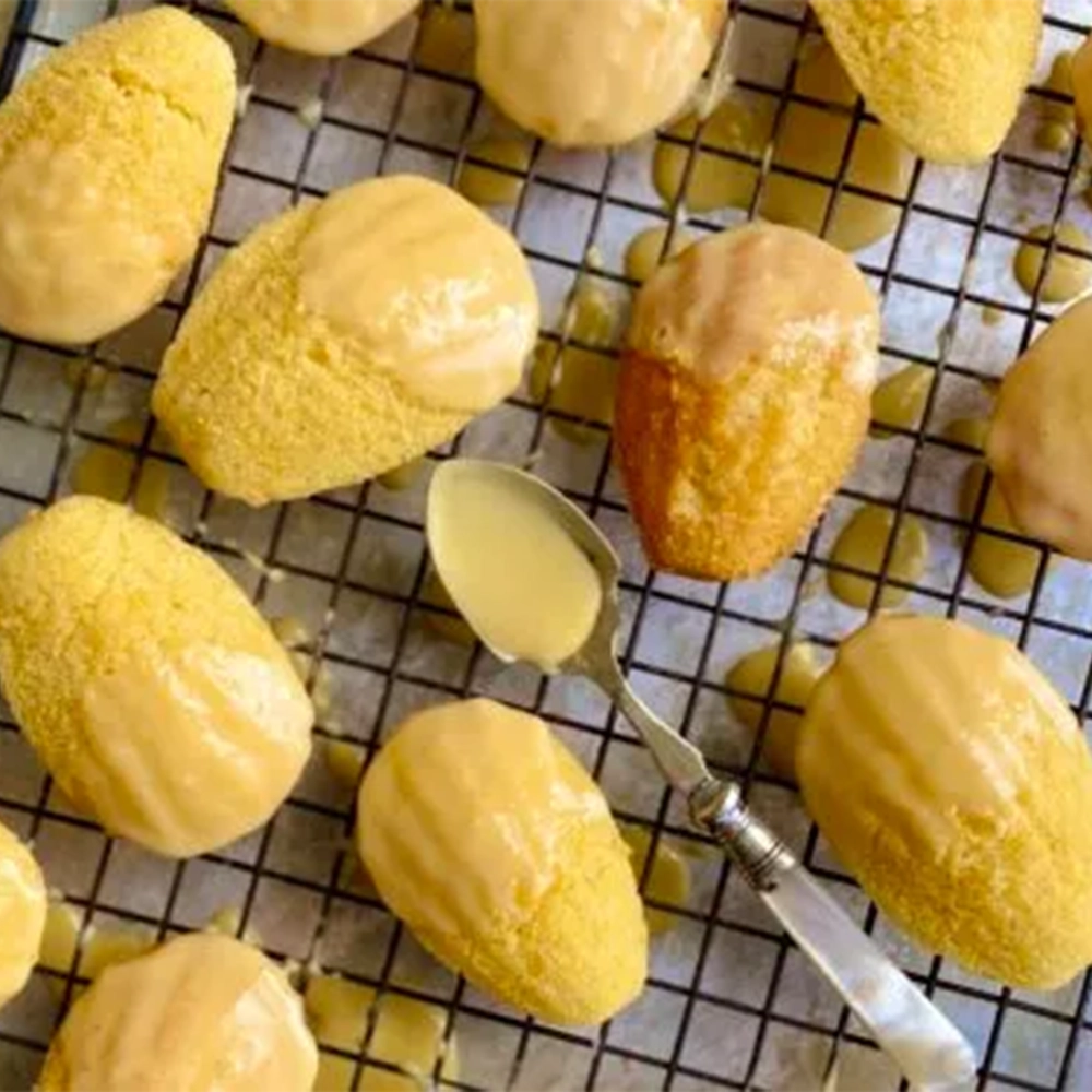 A baking tray with yellow passionfruit madeleines and a spoon liquid yellow icing sugar.