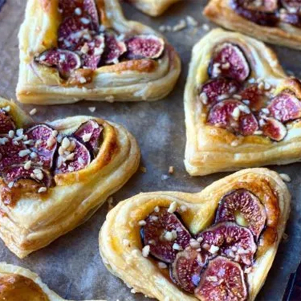 A baking tray with heart shaped butter puff pastries with thin red fig slices and chopped roasted hazelnuts sprinkled on top laid on baking paper.