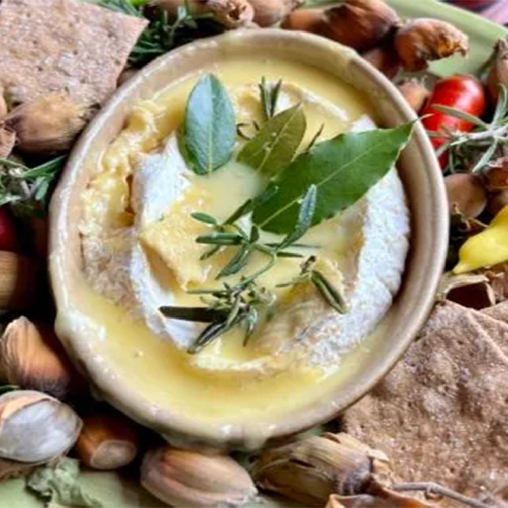 A small deep plate with a melted 'Baron Bigod' cheese with a bunch of herbs stuck inside the melded cheese surrounded by bread crackers, hazelnut, red and yellow chillies and rosemary twigs on a larger plate.