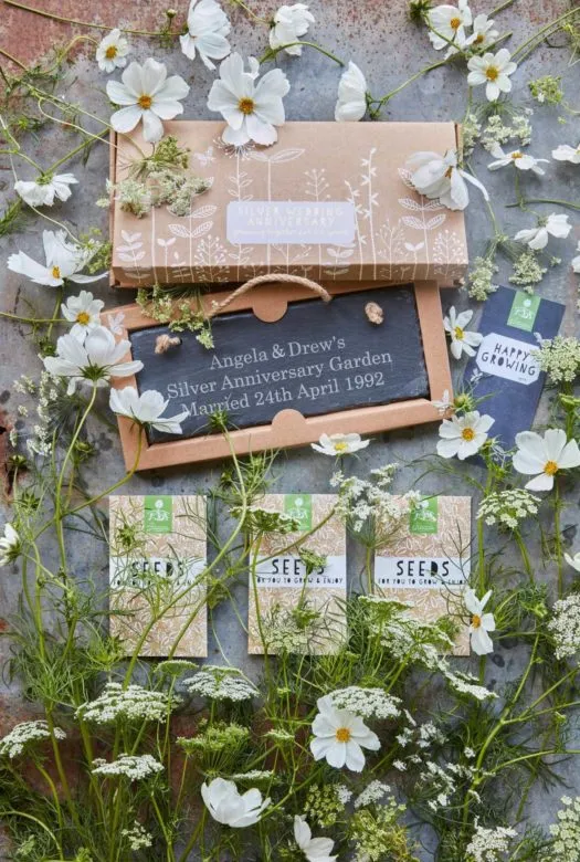 A decorated cardboard presentation box for a silver wedding anniversary with a selection of three packets of seeds, a large garden slate and a gift card, surrounded by white flowers of Lady's Lace, Cosmos Bipinnatus and White Beeblossom.