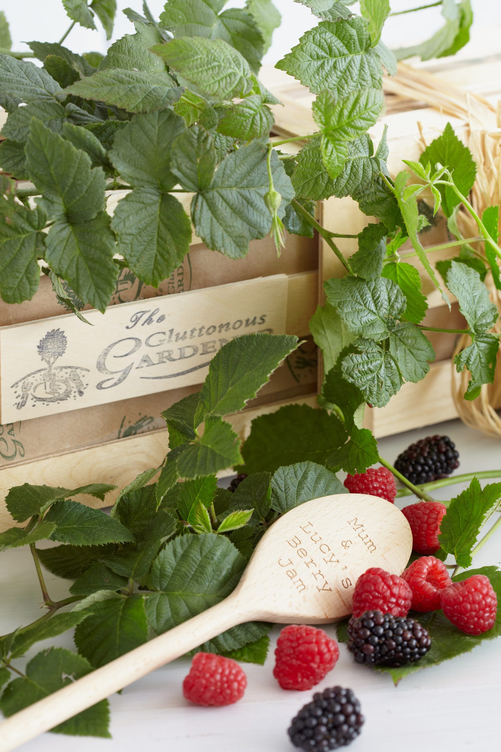 A personalised engraved wooden cooking spoon with a few fresh Loganberries and Blackberries nestled in the green foliage of a Loganberry Bush on a wooden gift crate.