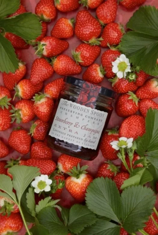 A jar of Strawberry & Champagne Extra Jam nestled in fresh strawberries, strawberry flowers and green strawberry leaves