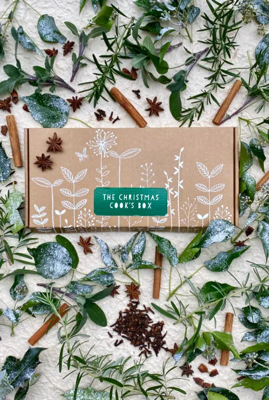 A small carboard box that reads 'Christmas cooks box' nestled in a collection of cinnamon sticks. rosemary twigs, dried star anise and cloves, twigs of thyme and other green leaves