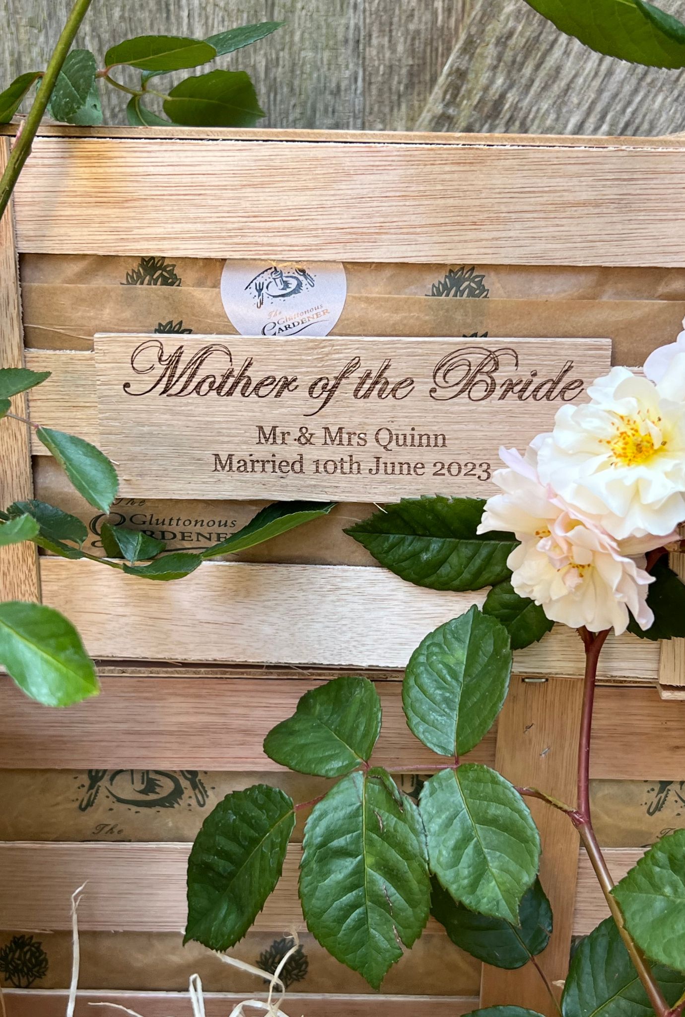 Close up of personalised Mother of the Bride wooden plaque on crate surrounded by leaves and white flowers.