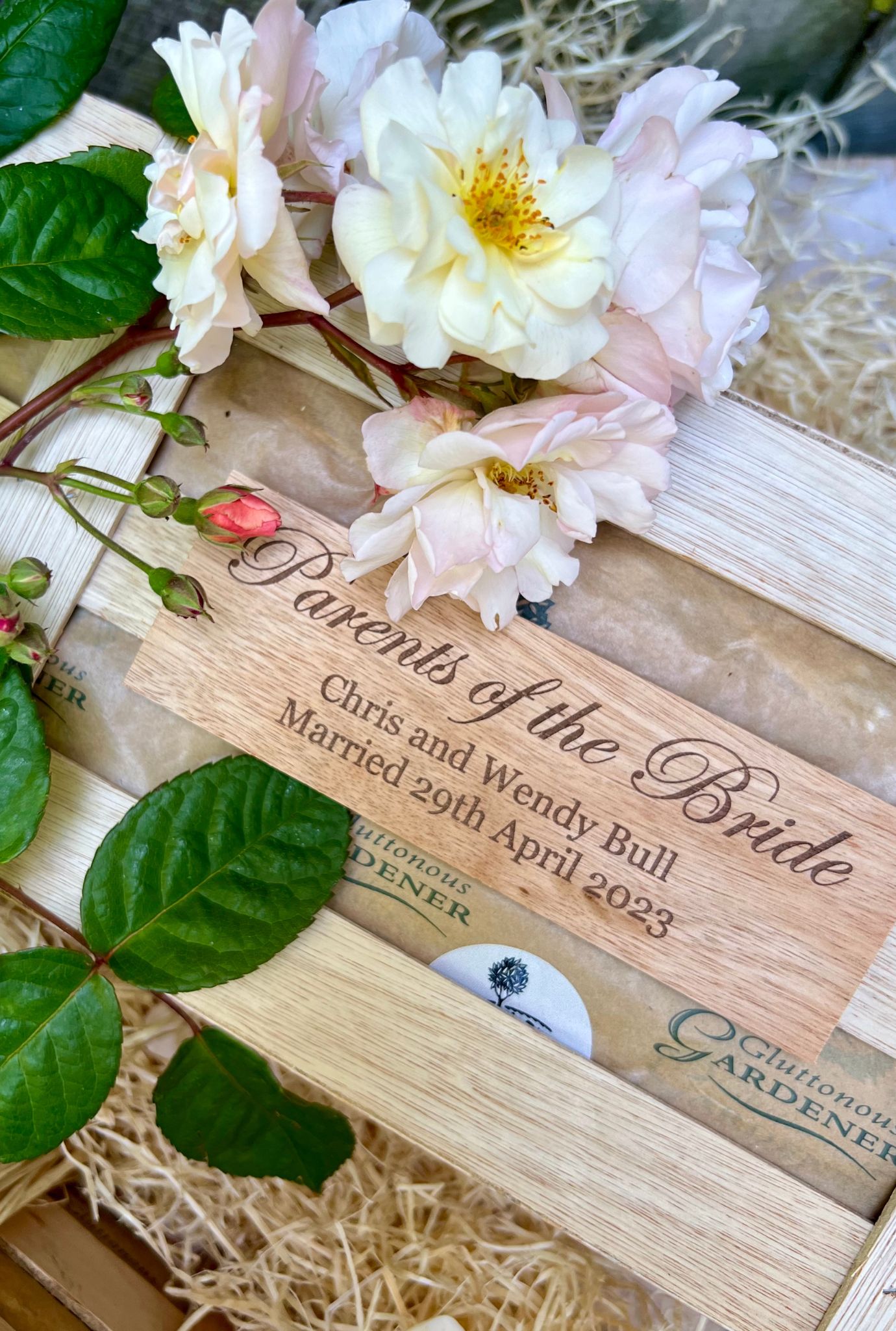 Close up of personalised wooden plaque on crate surrounded by rose plant leaves and pink flowers.