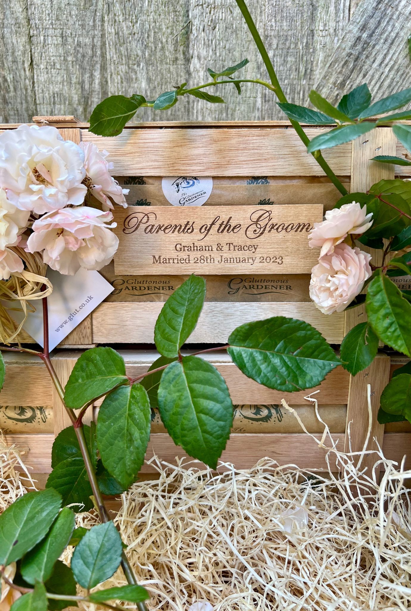 Close up of personalised wooden plaque on crate surrounded by rose plant leaves and pink flowers.