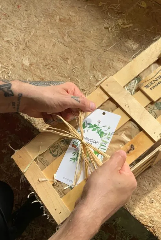 A wooden gift crate with an 'Anniversary' gift tag and two hands tying the ribbon.