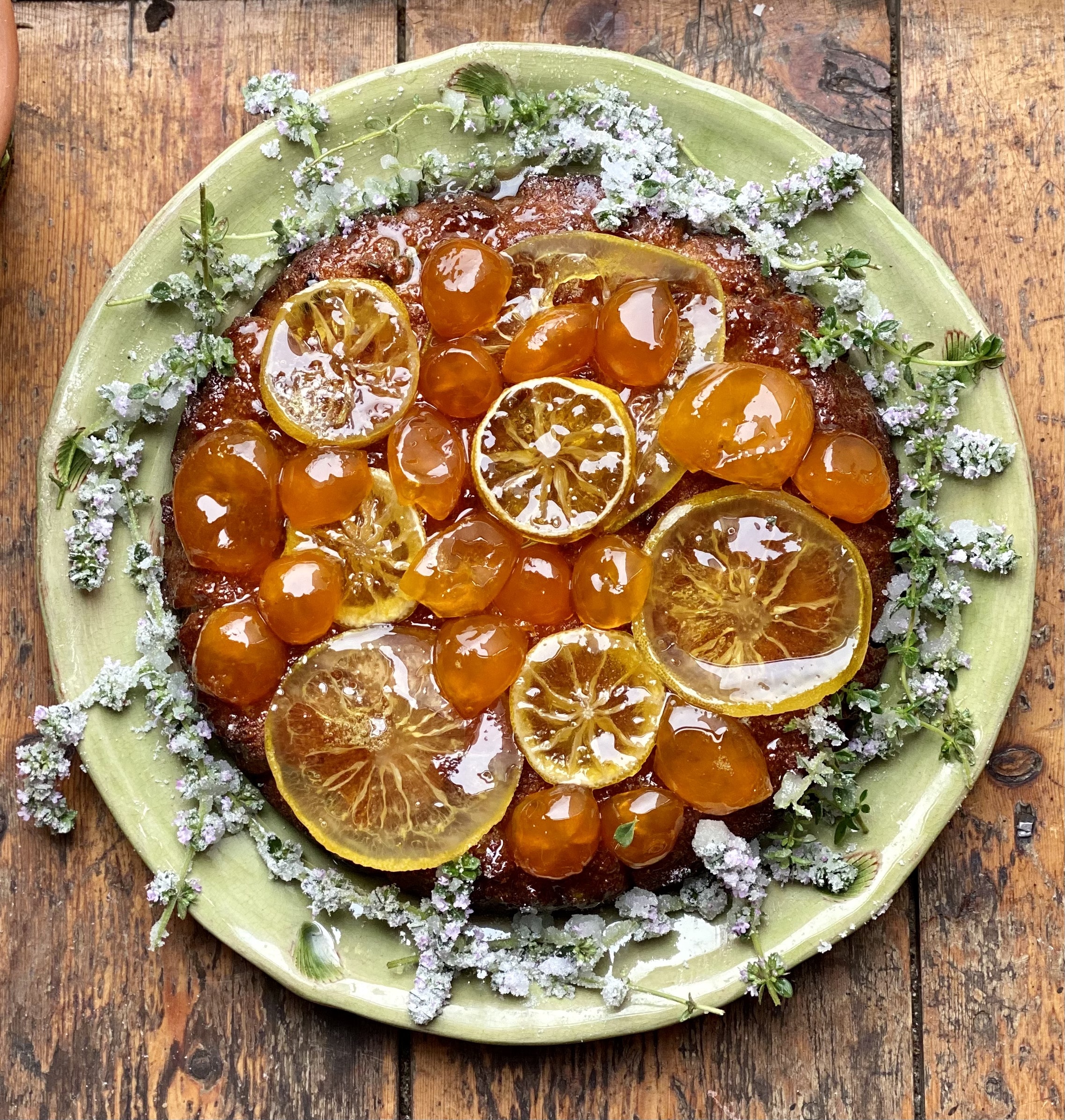 An orange fruit cake with jewel like sugared Calamondin oranges lemon slices on a green dish surrounded by sugared flowers.