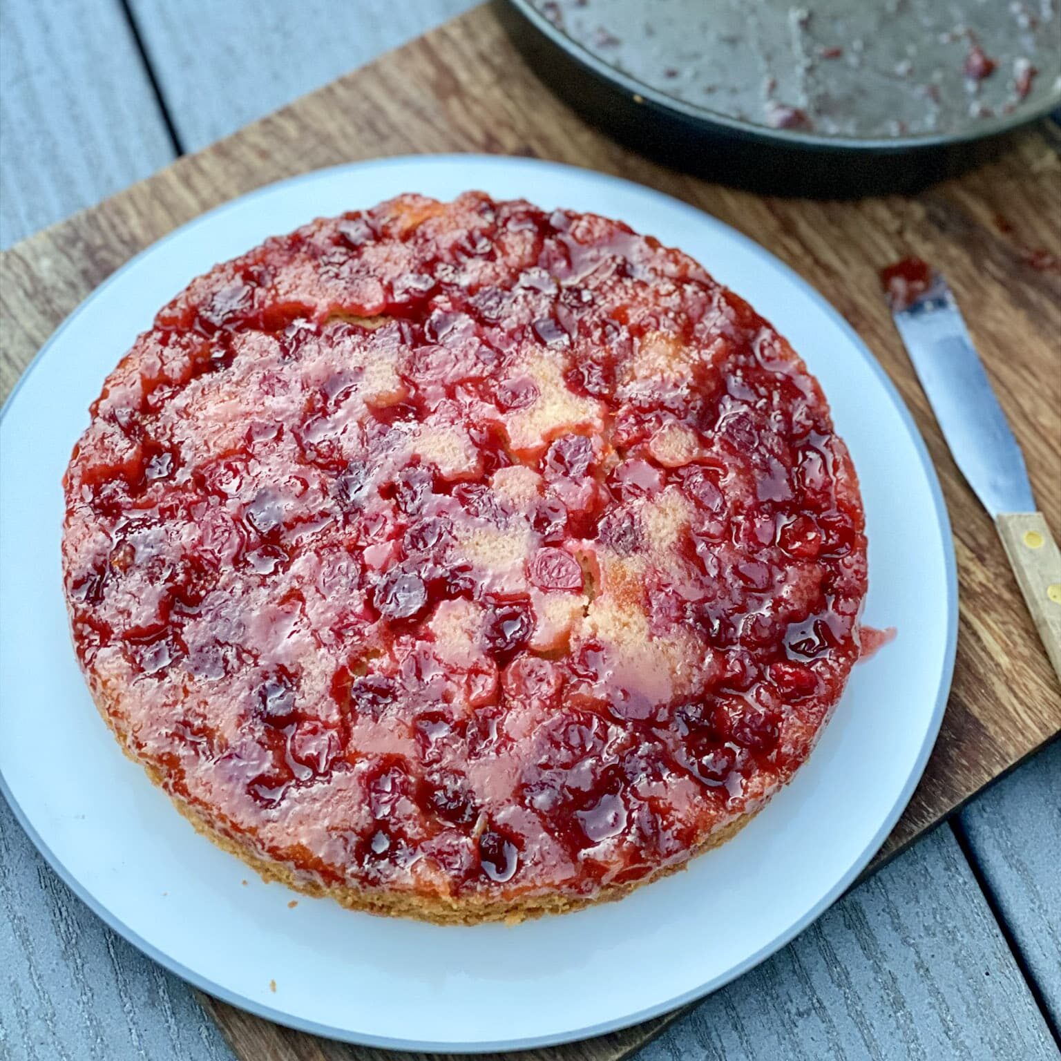 A cranberry cake with ruby red juicy cranberries on a white plate on top of a wooden board and a small knife.