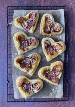 A baking tray with heart shaped butter puff pastries with thin red fig slices and chopped roasted hazelnuts sprinkled on top laid on baking paper.