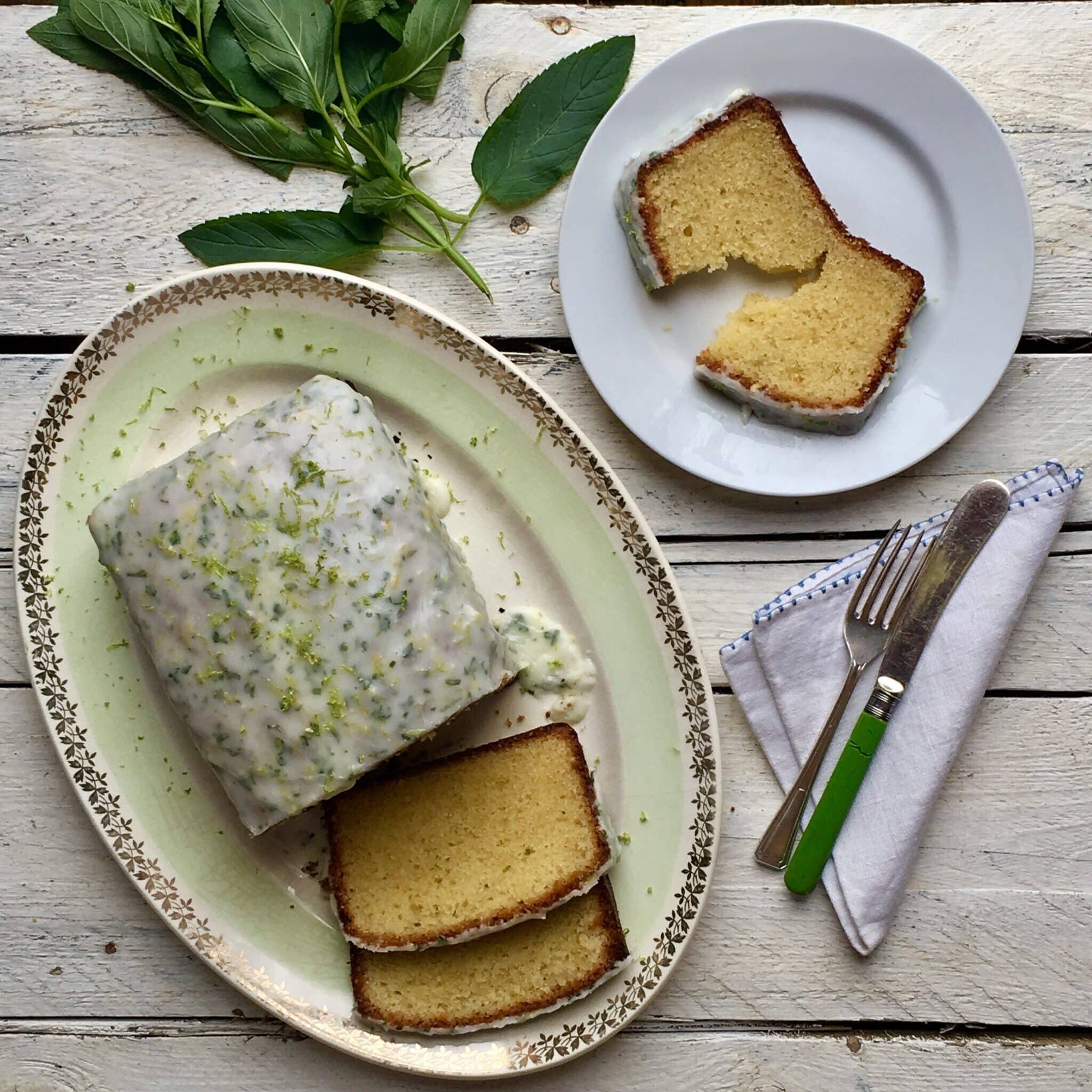 A sliced gin and lime loaf cake with a sugar frosting, lime zest and chopped peppermint leaves on a mint green decorative cake plate with a twig of peppermint leaves.