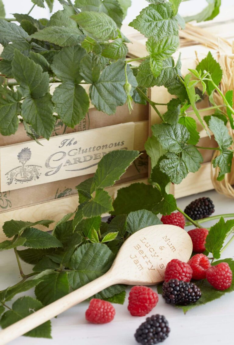 A personalised engraved wooden cooking spoon with a few fresh Loganberries and Blackberries nestled in the green foliage of a Loganberry Bush on a wooden gift crate.