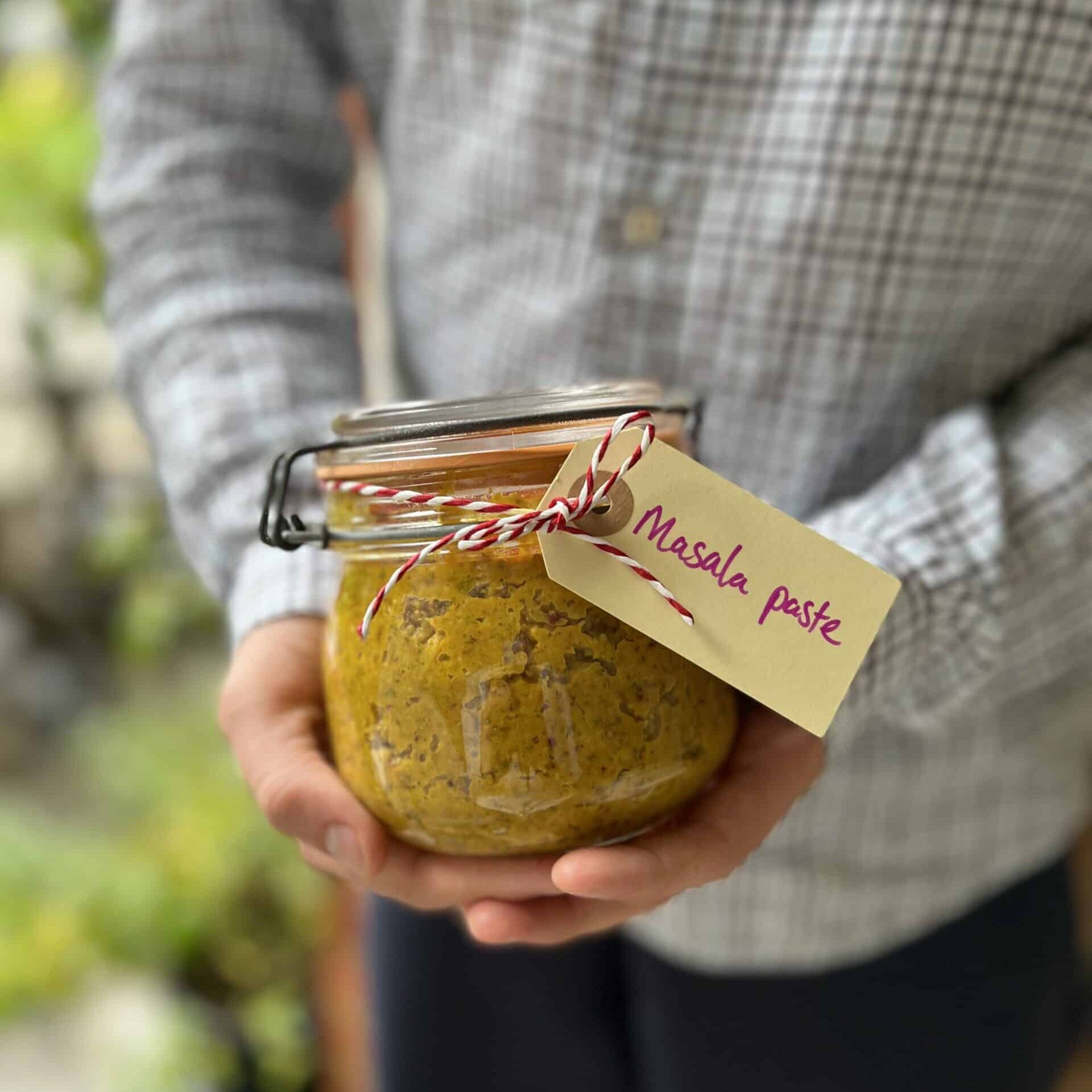 A canning jar with a beige - yellow Masala paste and a description tag held by two hands.