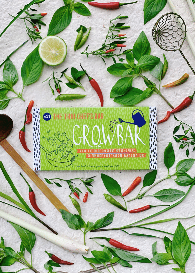 A green thai chef's growbar surrounded by thai basil leaves, birds eye chillies, a lemongrass stick. An open lime and a sieve.
