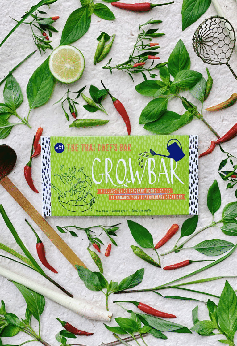A green thai chef's growbar surrounded by thai basil leaves, birds eye chillies, a lemongrass stick. An open lime and a sieve.