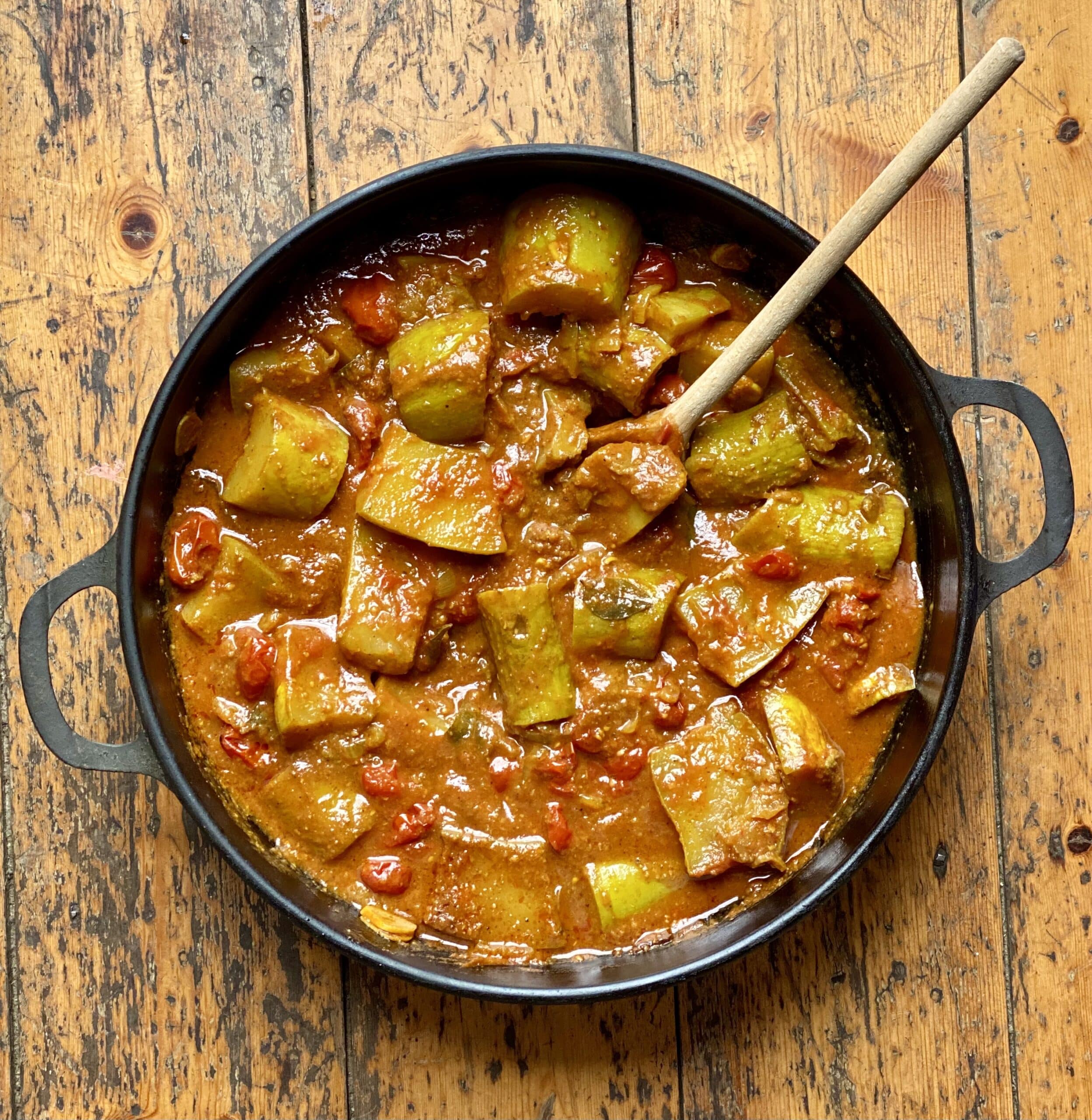 A cast iron pot filled with a Tromboncino masala meal with tromboncino squash, garlic and baby tomatoes in an orange coloured curry sauce.