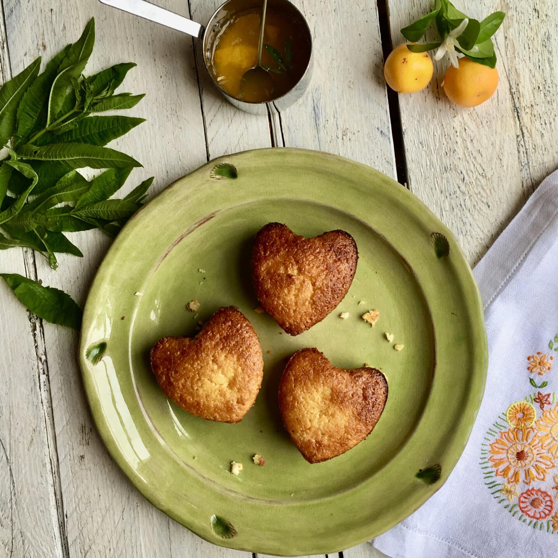 A green plate on a wooden table with three small heart shaped Calamondin Orange and Almond cakes next to a bunch of green leaves and to small yellow fruits.